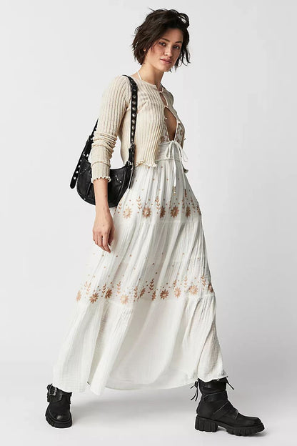 Woman wearing a long white maxi halter dress with floral embroidered patterns and black boots on a plain background