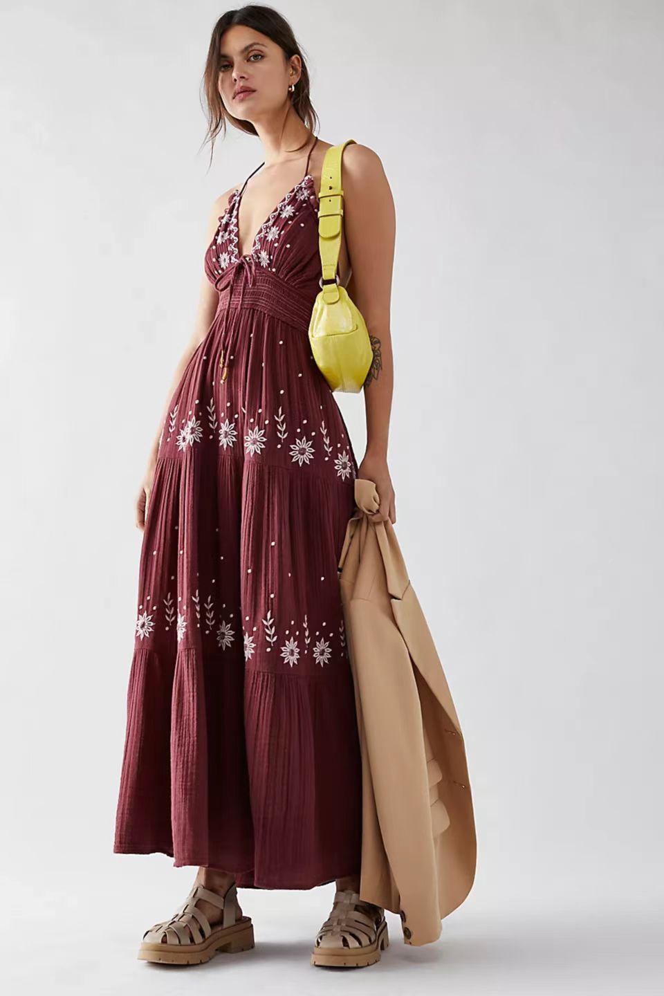 Woman wearing a burgundy embroidered maxi halter dress with a yellow bag and beige coat on a white background
